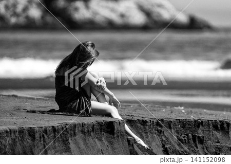 Woman sitting on cliff edge at beach. Lady looking down at sea. Retro style photo, grain effect Woman sitting on cliff edge at beach. Lady looking down at sea. Retro style photo, grain effect 141152098
