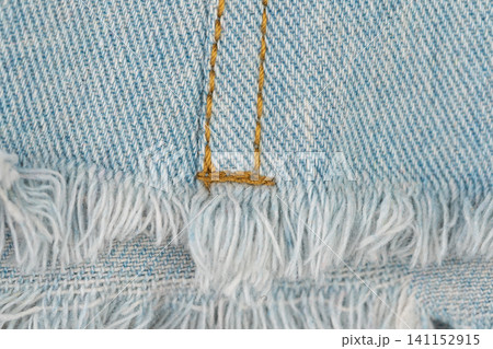 Close-up of light blue denim fabric with distressed fringe edge. Texture detail of casual jeans with detailed stitching. Trendy fashion material for textile background design 141152915