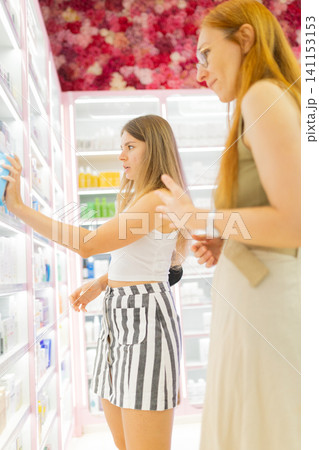 Two women shopping for skincare products in a brightly lit mall store with colorful shelves filled with various beauty items and floral decor in the background 141153153