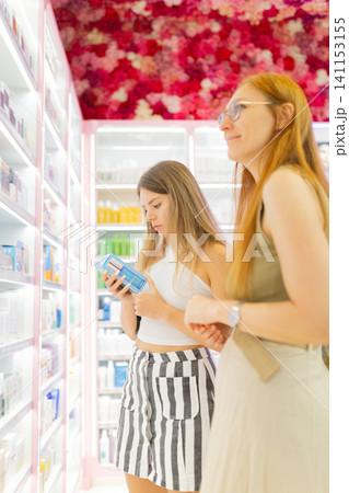 Two women shopping for skincare products in a brightly lit mall store with colorful shelves filled with various beauty items and floral decorations overhead 141153155