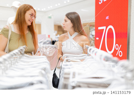 Two women shopping in a mall, browsing clothing racks with hangers, surrounded by a bright interior and a large sale sign displaying a discount 141153179