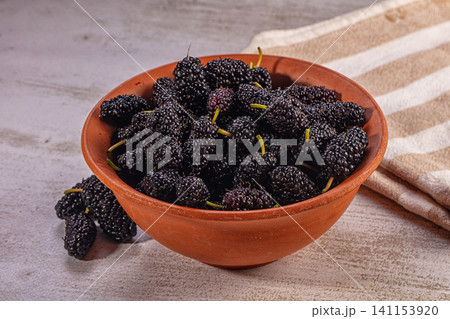 Fresh dark mulberries in rustic clay bowl on textured surface with striped cloth 141153920