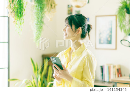 観葉植物に囲まれた部屋でスマホを操作する若い日本人女性 観葉植物に囲まれた部屋でスマホを操作する若い日本人女性 141154343
