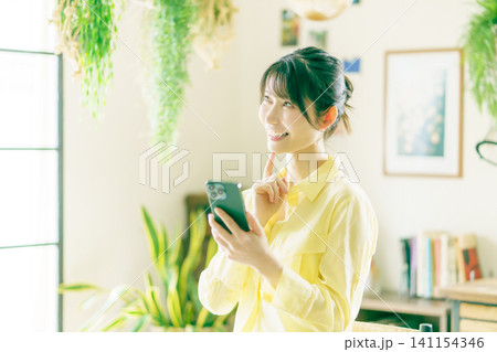 観葉植物に囲まれた部屋でスマホを操作する若い日本人女性 141154346