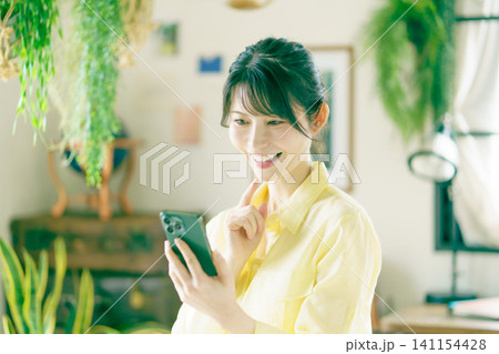 観葉植物に囲まれた部屋でスマホを操作する若い日本人女性 観葉植物に囲まれた部屋でスマホを操作する若い日本人女性 141154428