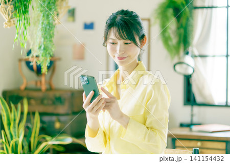 観葉植物に囲まれた部屋でスマホを操作する笑顔の若い日本人女性 観葉植物に囲まれた部屋でスマホを操作する笑顔の若い日本人女性 141154432