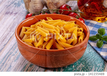 Uncooked penne pasta in rustic ceramic bowl with cherry tomatoes garlic basil and olive oil on weathered wooden table 141155489