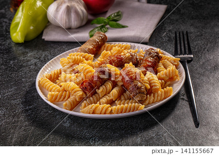 Fusilli pasta with bacon wrapped sausages on white plate with basil and garlic background Fusilli pasta with bacon wrapped sausages on white plate with basil and garlic background 141155667