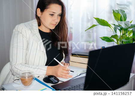 Young brunette woman sitting at her desk and using a pen to write down appointments and other notes in a notebook. Woman at her desk. Working from home.  141156257