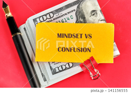 A note with the words mindset vs confusion sits on top of a stack of cash alongside a pen and a paperclip. The scene shows a workspace setup with financial elements A note with the words mindset vs confusion sits on top of a stack of cash alongside a pen and a paperclip. The scene shows a workspace setup with financial elements 141156555