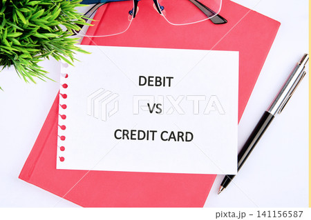 A simple view of a white paper with the words debit vs credit card on a pink notebook. A pen and glasses are also on the table along with a small green plant A simple view of a white paper with the words debit vs credit card on a pink notebook. A pen and glasses are also on the table along with a small green plant 141156587