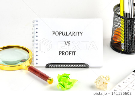 A notebook shows the words popularity versus profit. There are office supplies like a magnifying glass, crumpled papers, and colorful items on a white desk A notebook shows the words popularity versus profit. There are office supplies like a magnifying glass, crumpled papers, and colorful items on a white desk 141156602