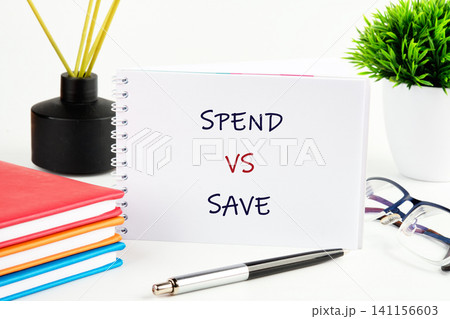 A notebook is open on a table showing spend versus save. There are colored notebooks, a pen, glasses, and a potted plant around it 141156603