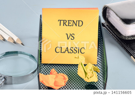 A sticky note with the words trend vs classic is placed on a black tray. Crumpled papers are around it along with pencils and a magnifying glass. Office supplies are in view 141156605