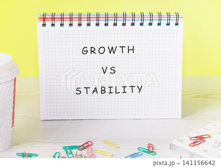 A notepad shows the words growth and stability side by side in large letters. Colorful paper clips lay on the desk near a cup. A bright yellow wall is in the background A notepad shows the words growth and stability side by side in large letters. Colorful paper clips lay on the desk near a cup. A bright yellow wall is in the background 141156642