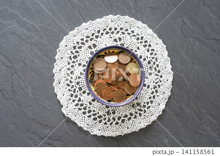 A bowl filled with euro coins sits on a white doily, which is placed on a dark, textured surface. A bowl filled with euro coins sits on a white doily, which is placed on a dark, textured surface. 141158561