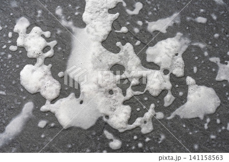 White foam with small bubbles spreads across a dark, textured surface. White foam with small bubbles spreads across a dark, textured surface. 141158563