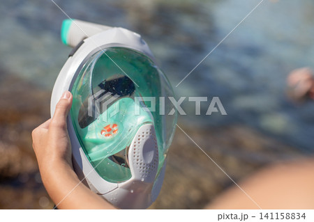 Person holding a full-face snorkel mask near clear water with rocky shoreline and blurred background of another individual swimming Person holding a full-face snorkel mask near clear water with rocky shoreline and blurred background of another individual swimming 141158834