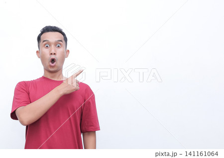Man in red t-shirt points with wide-eyed surprise. His mouth is open, showing shock or excitement. He gestures toward empty space, drawing viewer's attention. Perfect for advertising 141161064