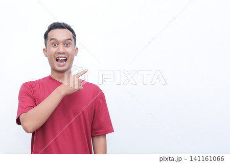 Man in red t-shirt points with wide-eyed surprise. His mouth is open, showing shock or excitement. He gestures toward empty space, drawing viewer's attention. Perfect for advertising Man in red t-shirt points with wide-eyed surprise. His mouth is open, showing shock or excitement. He gestures toward empty space, drawing viewer's attention. Perfect for advertising 141161066