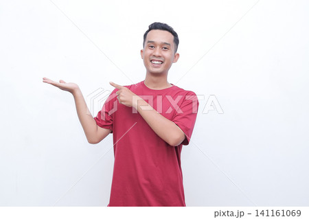 Young man smiles broadly, pointing with one hand. He wears a plain red t-shirt, standing against white. His open palm gestures toward an unseen product or idea 141161069
