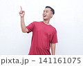 Young man gestures with joy pointing red t-shirt. He smiles warmly, conveying excitement or enthusiasm. Plain white background highlights his expression and motion.Ideal for advertising, presentations 141161075