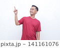 Young man gestures with joy pointing red t-shirt. He smiles warmly, conveying excitement or enthusiasm. Plain white background highlights his expression and motion.Ideal for advertising, presentations 141161076