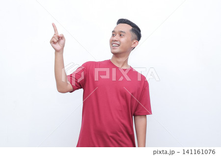Young man gestures with joy pointing red t-shirt. He smiles warmly, conveying excitement or enthusiasm. Plain white background highlights his expression and motion.Ideal for advertising, presentations 141161076