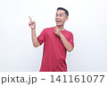 Young man gestures with joy pointing red t-shirt. He smiles warmly, conveying excitement or enthusiasm. Plain white background highlights his expression and motion.Ideal for advertising, presentations 141161077