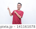 Young man gestures with joy pointing red t-shirt. He smiles warmly, conveying excitement or enthusiasm. Plain white background highlights his expression and motion.Ideal for advertising, presentations 141161078