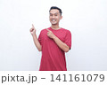 Young man gestures with joy pointing red t-shirt. He smiles warmly, conveying excitement or enthusiasm. Plain white background highlights his expression and motion.Ideal for advertising, presentations 141161079
