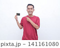 Young man smiles, pointing to credit card. He wears red t-shirt, holds card with pride. Neutral background keeps focus on his gesture. Image conveys confidence and approval 141161080