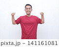 Young man smiles brightly, flexing arms. He wears a solid red t-shirt. His cheerful pose conveys confidence and energy. Ideal for fitness, wellness, or motivational themes 141161081