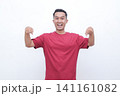 Young man smiles brightly, flexing arms. He wears a solid red t-shirt. His cheerful pose conveys confidence and energy. Ideal for fitness, wellness, or motivational themes 141161082