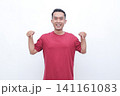 Young man smiles brightly, flexing arms. He wears a solid red t-shirt. His cheerful pose conveys confidence and energy. Ideal for fitness, wellness, or motivational themes 141161083