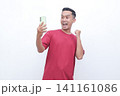 Man wears red shirt, holds phone, cheers with raised fist. Bright smile, eyes wide, exudes excitement and joy. Ideal for celebrating wins, success, or digital achievements 141161086