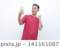 Man wears red shirt, holds phone, cheers with raised fist. Bright smile, eyes wide, exudes excitement and joy. Ideal for celebrating wins, success, or digital achievements 141161087