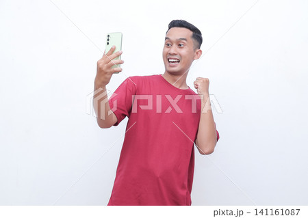 Man wears red shirt, holds phone, cheers with raised fist. Bright smile, eyes wide, exudes excitement and joy. Ideal for celebrating wins, success, or digital achievements Man wears red shirt, holds phone, cheers with raised fist. Bright smile, eyes wide, exudes excitement and joy. Ideal for celebrating wins, success, or digital achievements 141161087