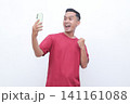Man wears red shirt, holds phone, cheers with raised fist. Bright smile, eyes wide, exudes excitement and joy. Ideal for celebrating wins, success, or digital achievements 141161088