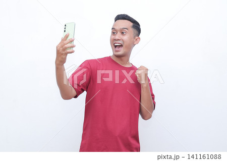 Man wears red shirt, holds phone, cheers with raised fist. Bright smile, eyes wide, exudes excitement and joy. Ideal for celebrating wins, success, or digital achievements Man wears red shirt, holds phone, cheers with raised fist. Bright smile, eyes wide, exudes excitement and joy. Ideal for celebrating wins, success, or digital achievements 141161088