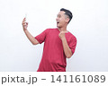 Man wears red shirt, holds phone, cheers with raised fist. Bright smile, eyes wide, exudes excitement and joy. Ideal for celebrating wins, success, or digital achievements 141161089