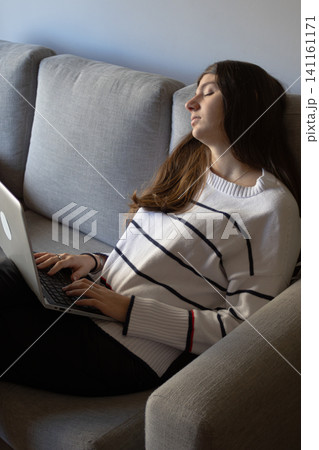 Young woman working remotely from her home, experiencing frustration and stress, attempting to work on her laptop while resting on a comfortable gray sofa Young woman working remotely from her home, experiencing frustration and stress, attempting to work on her laptop while resting on a comfortable gray sofa 141161171