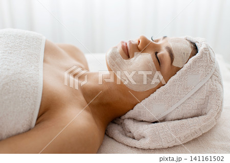 Black woman with facial mask applied, relaxing during skincare treatment at spa 141161502