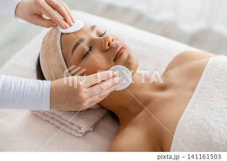 Cosmetologist cleansing face of black woman lying in spa with cotton pads Cosmetologist cleansing face of black woman lying in spa with cotton pads 141161503