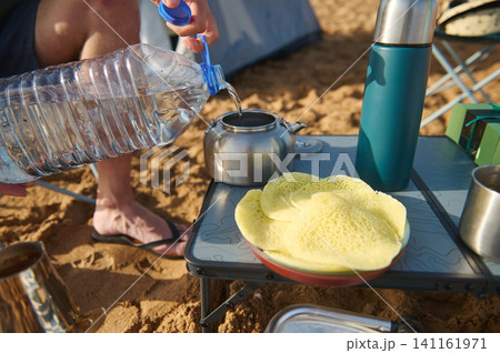 Camp breakfast on a sandy beach with pancakes, kettle and thermos as water is poured into a pot 141161971