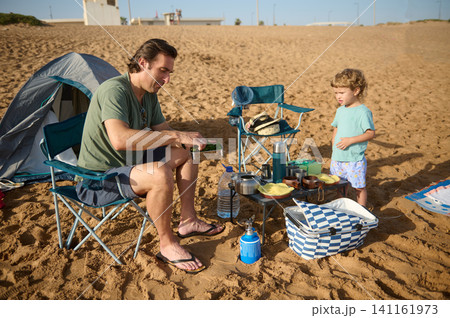 Father and young child camping on a sandy beach with picnic gear and portable stove 141161973