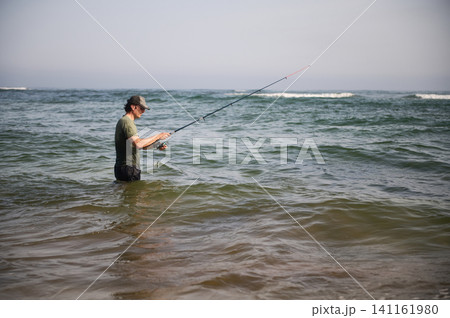Man fishing in the sea waist-deep casting a rod on a calm coastal day Man fishing in the sea waist-deep casting a rod on a calm coastal day 141161980