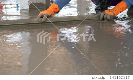 Mason worker working with trowel to level cement floor at construction site. A skilled construction worker spreading wet concrete to level wet concrete on a site floor. Foundation and flooring tasks. 141162117