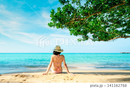 Woman in a pink bikini sitting on tropical beach facing turquoise ocean beneath a shady tree. Peaceful summer vacation, mindful relaxation, coastal wellness, travel lifestyle, and serene island escape Woman in a pink bikini sitting on tropical beach facing turquoise ocean beneath a shady tree. Peaceful summer vacation, mindful relaxation, coastal wellness, travel lifestyle, and serene island escape 141162788