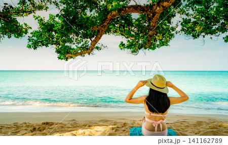 Back view of a woman wearing a straw hat relaxing beneath a tropical seaside tree. Digital detox, mindfulness, mental wellness, peaceful summer vacation, healthy lifestyle, and oceanfront tranquility. Back view of a woman wearing a straw hat relaxing beneath a tropical seaside tree. Digital detox, mindfulness, mental wellness, peaceful summer vacation, healthy lifestyle, and oceanfront tranquility. 141162789